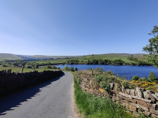 Dowry Reservoir, Holme, West Yorkshire. A narrow road bends down and to the left, with the reservoir on the right. Green hills in the background. 