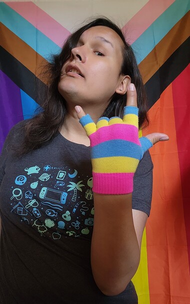 Me celebrating Pansexual Visibility Day (2023). I'm wearing my Pan gloves which the colors are yellow, light blue, and magenta; whilst making the punk hand gesture. I'm wearing my Animal Crossing t-shirt because why not. Background is the Pride Progress flag because (again), why not. 💗💛💙
