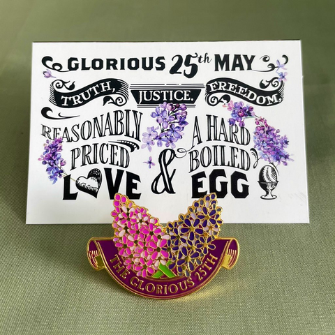 A white card with hand drawn sprigs of lilac between the words “Glorious 25th May” “Truth, Justice, Freedom, Reasonably Priced Love & A Hard Boiled Egg” sits on a sage green cloth. In front of the card, positioned so it does not obscure any of the writing, is a pin bag showing two crossed sprigs of lilac, one pink and one purple, inside a semicircular purple scroll with “The Glorious 25th” written in gold lettering.