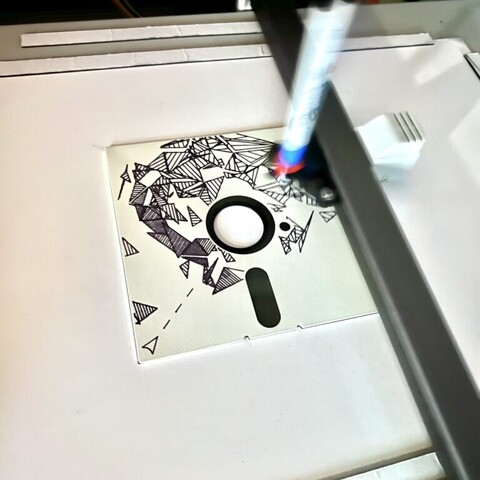 A picture of a #PenPlotter in action drawing on a white 5.25" floppy disk an abstract asteroid made up of triangles that is being disintegrated by a small triangular ship drawn in the left corner that is reminiscent of Asteroids the video game.