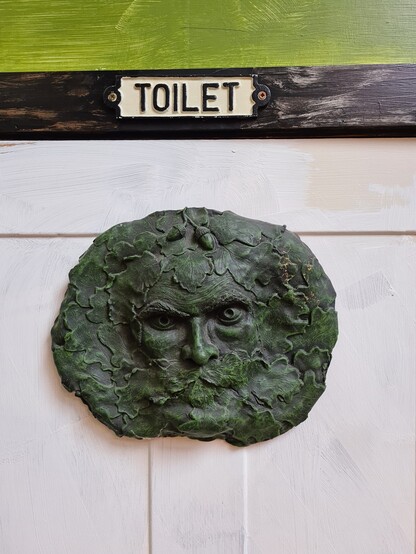 White wooden door with Toilet sign at top on the black wood door frame with green wall above with Green Man wood carving below at top of the door staring out at you from the picture