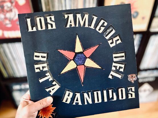 Hand holding a vinyl record (The Beta Band's Los Amigos del Beta Banditos) in front of a background of record shelves