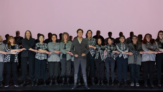 A group of women on a stage, all dressed in black trousers and white, grey or plaid flannel tops, standing in a row facing the audience, arms intertwined. Behind them, the silhouetted heads of men against a pale pink background. In the group on the left: soprano Megan Marie Hart as Tatyana in a red wig and glasses, in the middle: soprano Katrin Gerstenberger in riding boots as mother Larina, third from the right: mezzo-soprano Judith Christ-Küchenmeister as nanny Filippyevna.
Scene from Isabel Ostermann’s 2023 staging of Tchaikovsky’s opera Евгений Онегин (Eugen Onegin) at the Staatstheater Darmstadt. Photo © Nils Heck / Staatstheater Darmstadt
