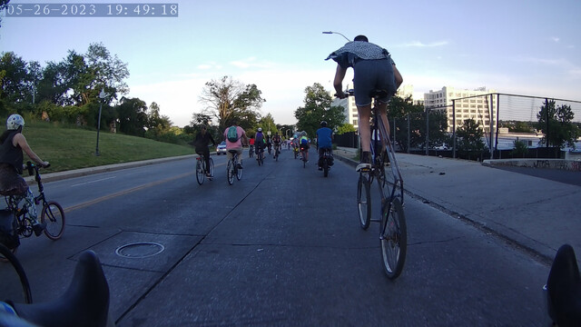 A stream of cyclists rolls down a steep hill.  A rider on a tall bike is visible in the foreground.