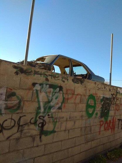 Tagged concrete brick wall of a scrap yard, crashed car, no doors, stacked high behind the wall. Clear blue sky. 