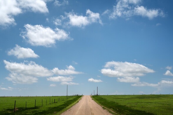 A dirt road meets the horizon, filled with blue skies and fluffy clouds.
