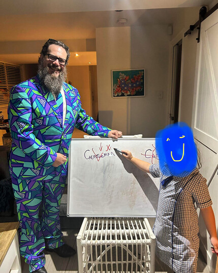 Photo of a bearded person wearing glasses and a neon (think late 80’s/90’s) three piece suit and a young child in school uniform. They are both facing the camera and smiling. They stand on either side of a whiteboard with the words “Gentleman” and “Cad” written on it. The child is pointing to the word “Gentleman”. The child’s face has been drawn over to hide their identity with a blue cloud and yellow smiley face drawn on it. 