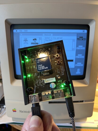 A picture of an AirTalk (a small circuit board in a plastic box with green LEDs) in front of a Mac Classic II showing the Chooser. A sticker on the AirTalk says Live, Laugh, LocalTalk from @billgoats