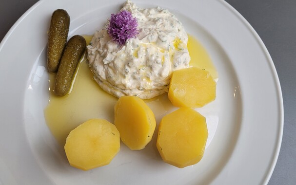 Auf einem Teller liegt eine Portion Kartoffeln mit Kräuterquark und Leinöl, im Quark steckt eine Schnittlauchblüte, daneben liegen zwei Cornichons. 