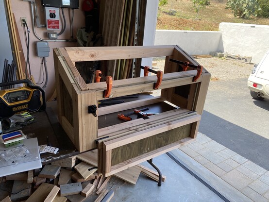 The nesting box in progress: a wooden box, unpainted, with a piece of trim still clamped in place. The box is sitting on a table in the garage.