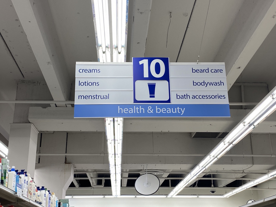Sign hanging over a pharmacy aisle that says “creams, lotions, menstrual, beard care, body wash, bath accessories”