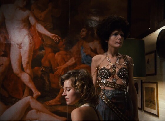 Still from the film The Bitter Tears of Petra von Kant, showing a seated young, blonde white woman with curly hair from the shoulders up in profile, with an older, middle-aged white woman standing behind her, thin and with black hair, in a skirt and a revealing top made up on strings of beadwork. They're both looking in different directions, pensive. Behind them an entire wall is covered in a reproduction of Poussin's Baroque painting Midas and Bacchus, naked bodies frozen, warm reds and pinks.