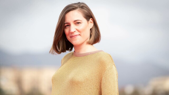 Foto en el exterior de Carla Simón, directora de cine. Mujer en la treintena, cara ovalada, leve sonrisa, rostro amable. Melena rubia, lisa con raya a la izquierda enseñando una oreja. Jersey mostaza, cuello redondo con  puntadas rosa. La ciudad difuminada al fondo.
