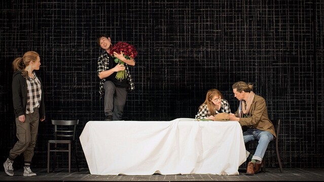 A man in a black and white plaid shirt, standing on a table covered with white cloth, holds a large bouquet of red roses. A red-haired woman in a black and white plaid shirt and a black jacket on the left is looking at him. On the right, a red-haired woman with glasses sits at the table and talks to a blond man at the right end of the table. He is wearing a light pink scarf, a brown jacket and blue jeans. Lena Sutor-Wernich as Olga, David Lee as Lensky, Megan Marie Hart as Tatyana, David Pichlmaier as Onegin.
Scene from Isabel Ostermann’s 2023 staging of Tchaikovsky’s opera Евгений Онегин (Eugene Onegin) at Staatstheater Darmstadt. Photo © Robert Schittko