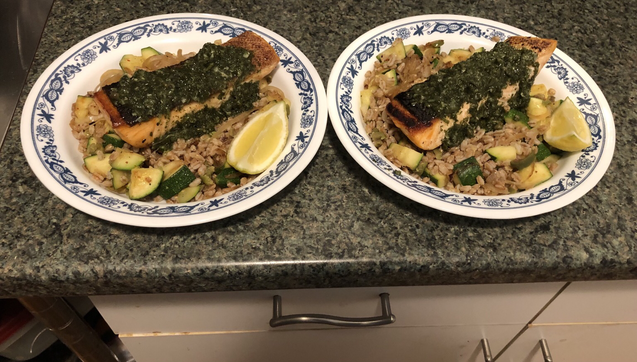 Plated dish. Trout on a bed of farro mixed with sautéed zucchini, onion, rehydrated golden raisins, and castelvetrano olives. Trout topped with Italian salsa verde. Lemon wedge on the side.