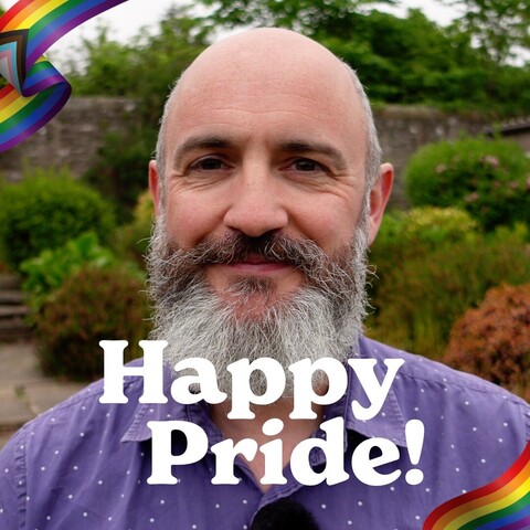 A selfie of me against an out of focus background of bushes, with ‘Happy Pride!’ overlaid on top in white.