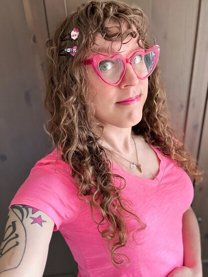 Me in a pink v-neck tee and fuchsia heart-shaped cat eye glasses. I have long curly brown hair and curly bangs, and a white/pink Monster High Skullita barrette and a pink heart barrette with “ghoul” on it in alternating black and white. I have dark eyeliner and bright pink lipstick that matches my shirt, pink stars and black lines from my tattoo are on my right bicep, and I am CUTE AF