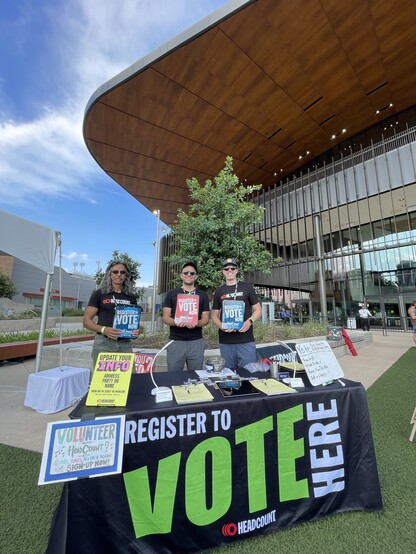 Three volunteers in Headcount shirts behind a Headcount table holding Headcount clipboards, in front of the Moody Center arena