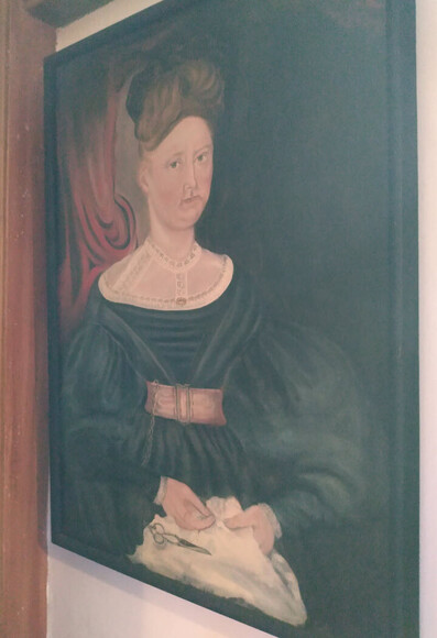 Apparently this is a portrait of Allethenia Fisk Ferrar, who is painted looking at the painter while holding a lap full of cloth and she appears to be sewing and has scissors attached to a chatelaine. I'd guess from the big poofy sleeves it's an 1830s outfit. 

More details here: https://www.historicnewengland.org/explore/collections-access/gusn/2753