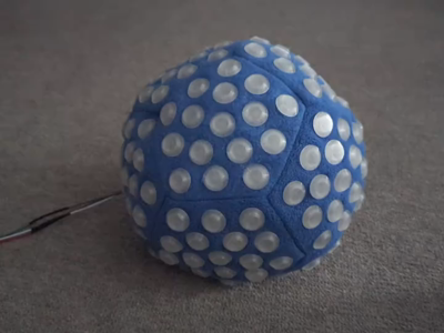 A soft blue fleece dodecahedron about the size of a soccer ball with a pattern of 16 translucent 3D printed diffusers on each side. The power cord extending from the zippered closure in the dodecahedron is plugged into a USB power pack and colorful patterns are displayed by the diffusers as the hands roll and squish the polyhedron.