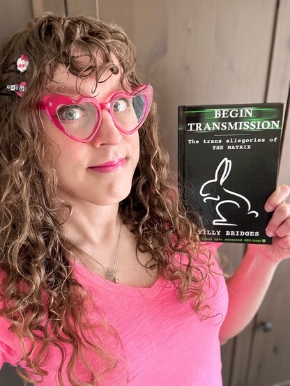 Me in a pink v-neck tee and fuchsia heart-shaped cat eye glasses. I have long curly brown hair and curly bangs, and a white/pink Monster High Skullette barrette and a pink heart barrette with “ghoul” on it in alternating black and white. I have dark eyeliner and bright pink lipstick that matches my shirt, I am CUTE AF, and I’m holding up a copy of my book, BEGIN TRANSMISSION: THE TRANS ALLEGORIES OF THE MATRIX