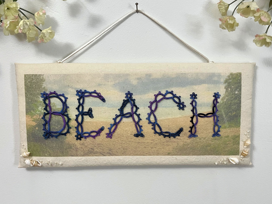 The word “Beach” spelled out in handmade lace letters on a background photo of a beach,