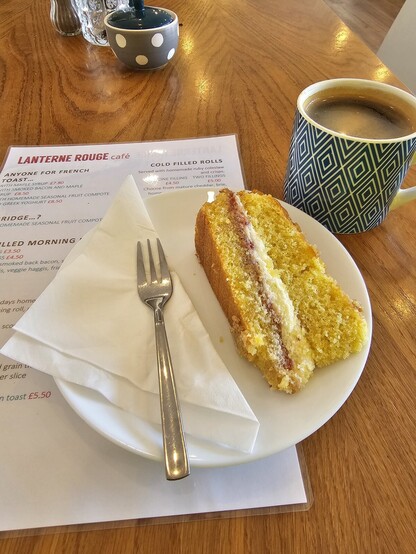 A slice of Victoria Sponge cake on a plate with a fork all on top of a cafe menu - alongside a cup of coffee
