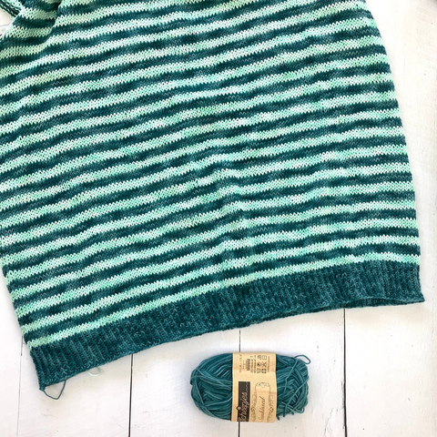 Flat lay view of the bottom half of a striped garment in teal and light turquoise. It is shown on a white plank background together with a ball of yarn.