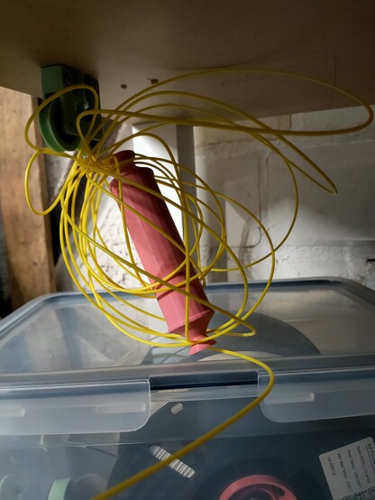 The underside of a table. Visible is a tangled knot of yellow filament with the axle of the filament holder entangled in it.
In the lower half of the image a diy drybox in which parts of another spool are visible.