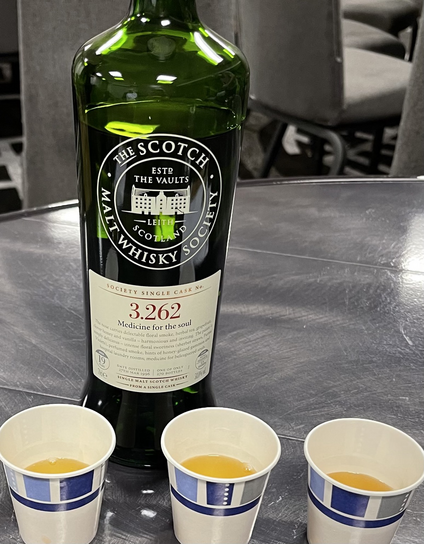A close up photo of a bottle of scotch. Three paper cups of scotch sitting in a row.