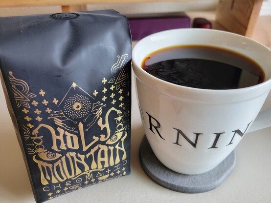 White 16-ounce mug of black coffee. The text wrapping around its circumference reads "Good morning". 

To its left is a 12 ounce pouch of coffee beans. The coffee pouch reads "Holy Mountain / Chromatic / 12 oz / 340 g / Mystical - Dark - Timeless".  The pouch has a black background with an intricate metallic gold design. The overall design may represent a mask. The design background features a grid of what look like cross fleury, "a cross adorned at the ends with flowers in heraldry." Thin gold lines and circles appear as connective elements of the design, perhaps representing pads and traces of an electronic circuit. At the top center appears a wide-open third eye surrounded by a geometric pattern of tiny circles (perhaps a lotus flower), all enclosed under an inverted V (perhaps representing a mountain). To its sides appear what might represent the horns of the mask, each made up of thin gold lines.  "Holy Mountain" appears below the third eye, in a font with funky, decorative brush strokes. Two snake heads extend from the characters of the word "Holy" with downward tongues touching an hourglass. Each hourglass, in turn, connects to a human eye with a spiral pupil. Centered below appears a t-shaped element which may represent a nose.  Below this appears the stylized name of the roaster, "Chromatic", with a few artistic flourishes. Below this appears two small half-bowls--one with text "12 oz", the other with "340 g". Below this appears the text "mystical / dark / timeless".