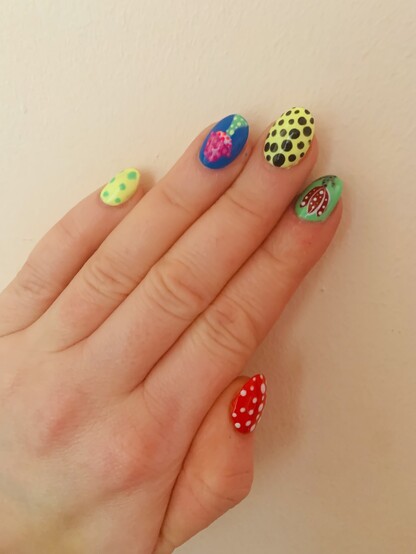 Nail art on the left hand. Left hand, starting from the pinky is bright yellow background and bright green spots, then bright blue back ground with pink/purple spotty and green toadstool, bright yellow background with black spots, red and white sporty Venus fly trap, and the thumb bright red with white spots.