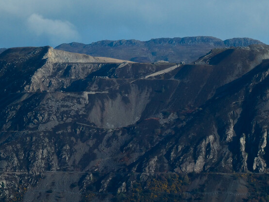 A view of Penmaenmawr granite quarry taken from seaward. The mountainside is gouged with the levels and terraces of the workings, with some areas of loose scree. Some bright flashes of yellow gorse and rust-coloured bracken break up the otherwise drab colours whilst the clefts are in heavy shadow. The ridge of the mountain range beyond is visible in the distance.
