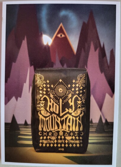 3"x5" postcard (front), showing a 12 ounce bag of Holy Mountain coffee in front of a painting of stylized, craggy mountains in various shades of purple, as seen in the distance from across a plain scattered with smaller pyramidical structures (trees, perhaps). A golden, winding path leads from the viewer to the mountains. A triangular mountain in the center is red with gold border, with a solitary eye at its peak. The sun is rising / setting just behind it. The bag of Holy Mountain coffee is black with an intricate gold design, possibly resembling a mask with a third eye. It has text Holy Mountain / Chromatic / 12 oz / 340 g / mystical - dark - timeless / HVG (seems to represent Hiver van Geenhoven, Head Roaster & Green Buyer)