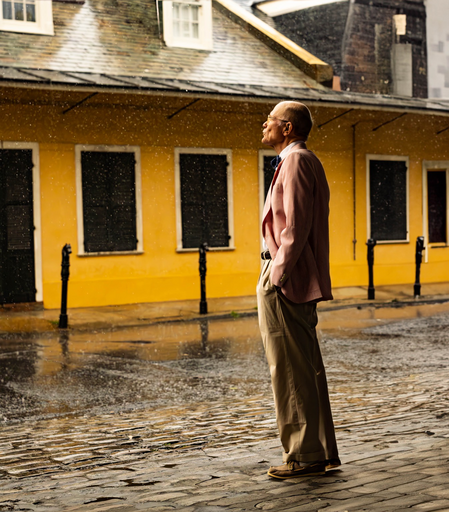 Man stands off right amongst a New Orleans street. It is raining. He is wearing pants a black belt and a pinkish blazer, also a bow tie!