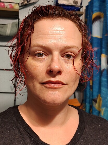 Photo of a white woman in her mid 40s, Brown eyes, brown and pink hair that's wet and thin and curly. She's looking directly into the camera with a slight smile on her face.