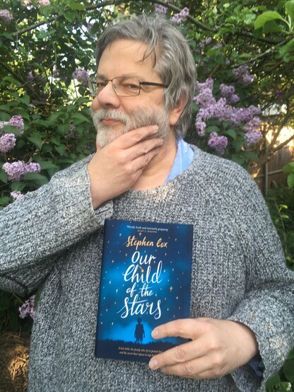 Stephen Cox Author - grey hair, beard and sweater - poses with his novel stroking bread - novel is Our Child of the Stars - background is a blooming lilac