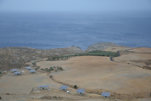 Das Foto zeigt eine steinige, karge,  Küstenlandschaft von einem Berg herab aufgenommen. In der oberen Hälfte des Fotos sieht man das blaue Meer. Es ist leicht dunstig und der Horizont geht in den Himmel über. Die abschüssige Landschaft  in der unteren Hälfte des Fotos ist vereinzelt mit kleinen Olivenhainen durchsetzt. Acht kleine Solaranlagen fügen sich in die Landschaft ein.
