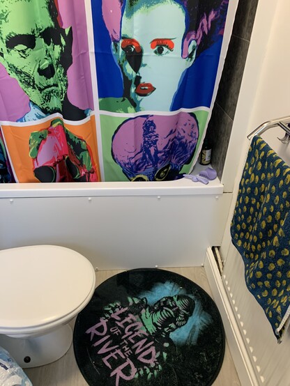 White bathroom suite, Shower curtain with a white background, with 9 universal monsters in an Andy Warhol style. Top left is Frankenstein’s monster in pink, blue, black on a yellow square background.  Dracula is blue, yellow, black on red square background. The wolf man is orange, yellow, green, black on a purple square background. The creature is oranges, yellow, purple on green square background. The mummy is green, blue, red on a purple Square background. Frankenstein’s bride is blue, red, purple, black, green on a blue square background. The phantom is blues, black on a square green and purple background. The invisible man is pinks, green, black on an orange background. Metaluna is purples, blue, yellow, black on green background. Round black bath mat on the floor, with a green creature from the black lagoon and legend of the river in purple lettering. Green and yellow leaopard print towel on the handrail.
