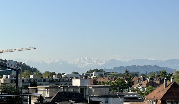 Der Himmel gen Süden ist eher hellblau und noch leicht dunstig. Von links ragt der Ausleger eines Bau-Krans ins Bild. Über den Häusern von Ostermundigen und grünen Rücken des Belpbergs sieht man die schneebedeckten Berner Alpen