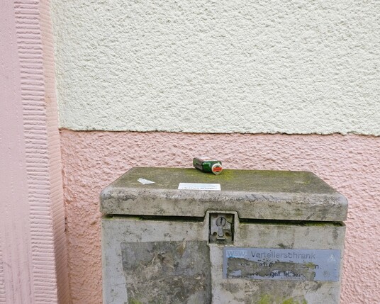 Foto einer liegenden Kräuterlikörflasche  auf einem Verteilerschrank vor einer rosa-weißen Hauswand. 