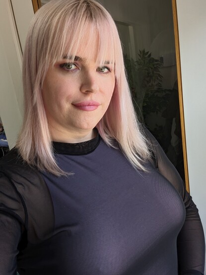 Selfie of a woman with very light pink hair in a lob with bangs, and purple eyeshadow and lipstick, wearing a sleeveless lilac bodysuit under a long-sleeved sheer top.
