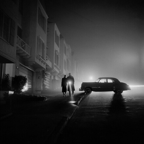 One of Brassaï’s night photos, of a car and two people backlit on a quiet street.