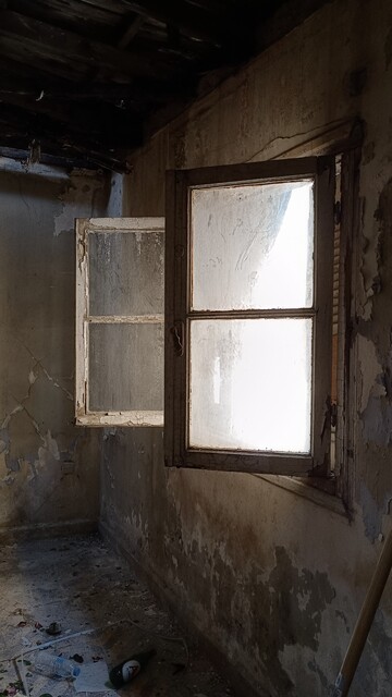Abandoned house, interior, side view of a wall with a casement window. Sashes are open, looking like two E's. A beer on the ground, among other rubbish. The ruined roof and a couple of buff cockroaches prevented me from stepping further inside. 