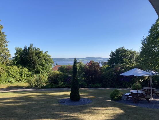 View over the Tay from a hotel garden in the sun, a narrow evergreen tree is in the centre and a bar table with parasol to the right 