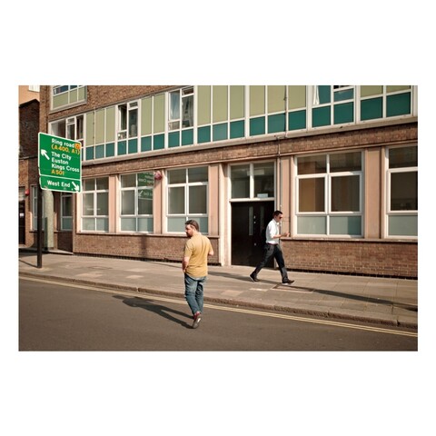 A modernist block in strong sunlight. Two people are in front of the building, one walking past, the other crossing the road.