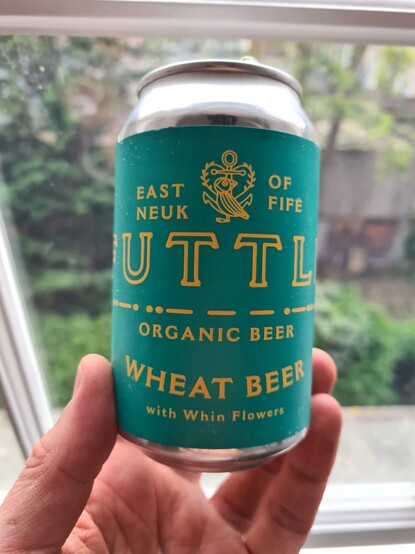 Hand holding up a silver beer can covered in a turquoise label with gold writing saying Futtle organic wheat beer behind beer can is out of focus window pane with white wood and an out of focus garden behind it