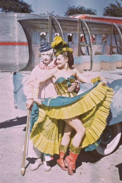 A clown with arm around waist of a dancer by a circus trailer 
