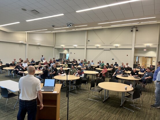 A large room filled with people sitting at tables listening to the opening session at Iowa Code Camp.