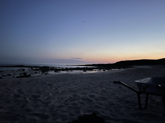 A scene of a beach and headland. Behind the headland the sky is slightly orange with the rest of it a milky blue. You can clearly see stuff on the beach and rocks sticking up. The photo was taken at half past midnight. 